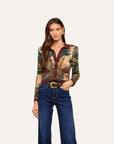 A woman modeling a long-sleeve button-up shirt with a detailed tapestry-style print in green, beige, and brown, paired with dark blue jeans and a gold-buckled belt, against a solid off-white background.
