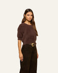 A woman modeling a short-sleeve brown knit top with a crew neck, paired with dark brown wide-leg trousers and a gold buckle belt, against a solid off-white background.