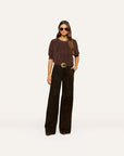 A woman modeling a dark brown short-sleeve knit top and dark brown wide-leg pants with a gold-tone belt, standing against a solid off-white background.
