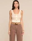 A woman modeling a cream-colored textured knit tank top with three gold buttons, paired with brown wide-leg jeans and a brown belt, against a solid beige background.