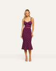 A woman modeling a sleeveless midi dress with a sweetheart neckline, thin shoulder straps, a fitted bodice, and a flared skirt. The dress is deep purple with a white polka-dot pattern and a thin purple waistband.