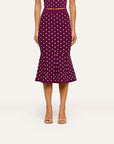 A model wearing a dark purple midi skirt with a white polka dot pattern, paired with a matching crop top and beige heeled sandals, set against a plain light background.