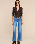 A model wearing a sheer black long-sleeve button-up shirt, blue flare jeans with a brown belt, and brown ankle boots, standing against a solid beige background.