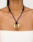 A close-up of a model wearing a black cord necklace with a polished gold circular pendant.