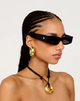 A close-up of a model wearing black rectangular sunglasses with a small gold star detail on the temple, along with a black cord necklace and gold hoop earrings, against a light gray background.