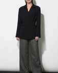 A model stands against a white studio wall wearing a black belted wrap blazer with wide long sleeves, paired with wide-leg charcoal gray trousers and black ankle boots.