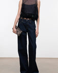 A model wearing a black sleeveless sheer mesh top with feather trim, paired with dark blue wide-leg jeans and holding a small patterned clutch, set against a plain white studio background.