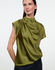 A woman modeling a sleeveless olive green satin top with a draped high neck, paired with dark blue jeans, against a plain light gray background.