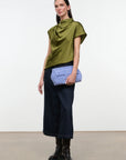 A model stands against a plain white studio wall wearing a draped olive green short-sleeve top, dark navy wide-leg cropped pants, black leather ankle boots, and holding a light blue envelope-style clutch.