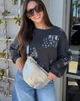 Woman wearing a grey sweatshirt with graphics, blue jeans, and white sneakers, holding a silver handbag.