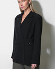 A woman modeling a black wrap-style blazer with a self-tie belt and notched lapels, paired with gray trousers, against a plain light gray wall.