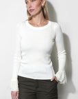 A woman modeling a white long-sleeve crewneck knit sweater with ruffled cuffs, paired with dark olive shorts and black tights, standing against a plain light gray wall.