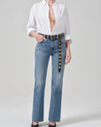 A model wearing a white button-down shirt, light-wash blue jeans with a decorative black belt featuring gold-tone circular hardware, and black pointed-toe heels, photographed against a light gray studio background.