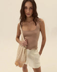 Woman wearing a beige tank top and white shorts, holding a mesh bag on a plain background