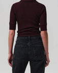 Person wearing a dark brown polo shirt and black jeans on a light gray background back view