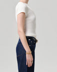 Side profile of a model wearing a white short-sleeve ribbed knit top tucked into dark blue jeans with a black belt, accessorized with a gold chain bracelet and ring, against a light gray studio background.