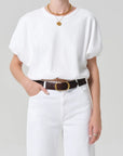 Person wearing a white top and white pants with a brown belt on a plain background