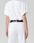 Person wearing a white blouse and white pants with a black belt on a plain background