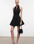 A model stands against a plain white studio wall wearing a black halter-neck mini dress with a flared skirt and holding a small black feathered handbag.