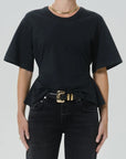A model wearing a black short-sleeve crewneck t-shirt with a peplum hem, styled with a black belt featuring a prominent gold buckle, paired with dark jeans and a gold chain necklace.