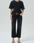 A model wearing a black short-sleeve crewneck t-shirt, black cropped wide-leg jeans with a gold-tone belt, and black pointed-toe heels against a light gray studio background.