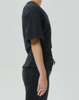 Side profile of a person wearing a black short-sleeve t-shirt and black jeans against a light gray studio background.