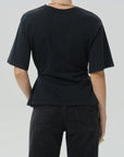 Rear view of a person wearing a black short-sleeve crew-neck T-shirt and dark jeans against a light gray studio background.