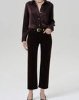 A model wearing a dark brown satin long-sleeve button-up shirt and matching dark brown velvet cropped trousers, styled with a gold belt, necklace, and black pointed-toe heels against a light gray studio background.
