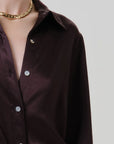 Close-up of a person wearing a dark brown satin button-up shirt with silver buttons and a chunky gold chain necklace, against a light gray background.