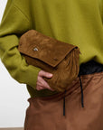 A person wearing an olive green sweater and brown skirt holds a brown suede flap pouch bag with a silver-tone metal clasp.
