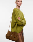 A model wearing an olive green knit sweater and brown pants, carrying a brown suede shoulder bag with a small silver clasp against a plain light gray background.