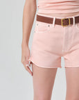 Person wearing pink shorts and a light pink top with a brown belt on a light gray background