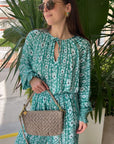 Woman in a green floral set holding a woven handbag, standing outdoors with plants in the background.