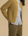A woman modeling a mustard yellow ribbed knit cardigan over a blue and white striped button-down shirt and olive green pants, set against a solid beige studio background.