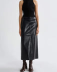 A model wearing a black sleeveless knit top and a black faux-leather midi skirt, paired with dark brown heeled ankle boots, against a light gray studio background.