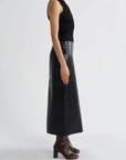 A model shown from the side wearing a black sleeveless knit top and a long black faux-leather midi skirt with a high waist, paired with dark brown heeled ankle boots, against a light gray studio background.