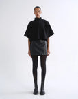 A model stands against a plain white studio background wearing a black short-sleeve mock-neck cape top, a black textured mini skirt, black tights, and black ankle boots.