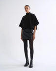 A model stands against a plain white studio background wearing a black high-neck cape-sleeve top, a black mini skirt with tonal side panels, black tights, and black ankle boots.