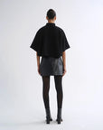 A model viewed from the back wearing a black short-sleeved high-neck top, a textured black mini skirt, black tights, and black ankle boots, set against a plain light gray studio background.