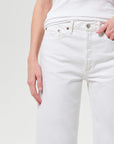 Close-up of a person wearing white jeans with a white ribbed top, showing the front waistband, belt loops, front pockets, and hands at the sides, against a light gray studio background.