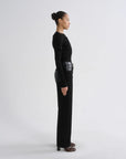 A model in profile wearing a black long-sleeve knit top and matching wide-leg trousers, styled with brown heeled shoes, against a plain light gray studio background.