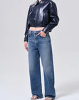 A model wearing a shiny dark navy cropped leather jacket and wide-leg blue jeans, standing against a light gray studio background.