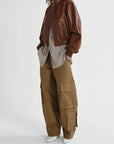 A model wearing a brown leather bomber jacket, a light gray shirt, brown cargo pants, and white pointed shoes, photographed against a plain light gray studio background.