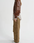 A model shown from the side wearing a brown faux-leather jacket, a light beige shirt, and wide-leg brown cargo pants against a plain light gray background.
