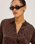 Woman wearing a brown polka dot shirt with sunglasses on a plain background