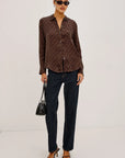 A woman modeling a brown long-sleeve button-up shirt with a small black polka dot pattern, paired with dark blue jeans, black sunglasses, and black heeled sandals, holding a black handbag against a plain white studio background.