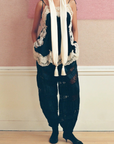 A model stands against a pink and cream wall wearing a black lace-trimmed camisole vest, navy and black lace pants, and a long cream scarf.