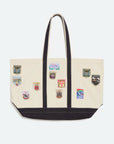 An off-white canvas tote bag with black trim, dual shoulder straps, and multiple colorful embroidered sports crests on the front, shown against a light gray background.