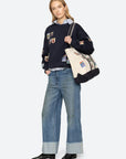 Person wearing a navy sweater with patches and blue jeans, holding a beige tote bag on a white background