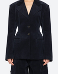 A model wearing a dark navy blue corduroy blazer with structured shoulders and a double-breasted button closure, paired with matching dark pants, against a light gray studio background.